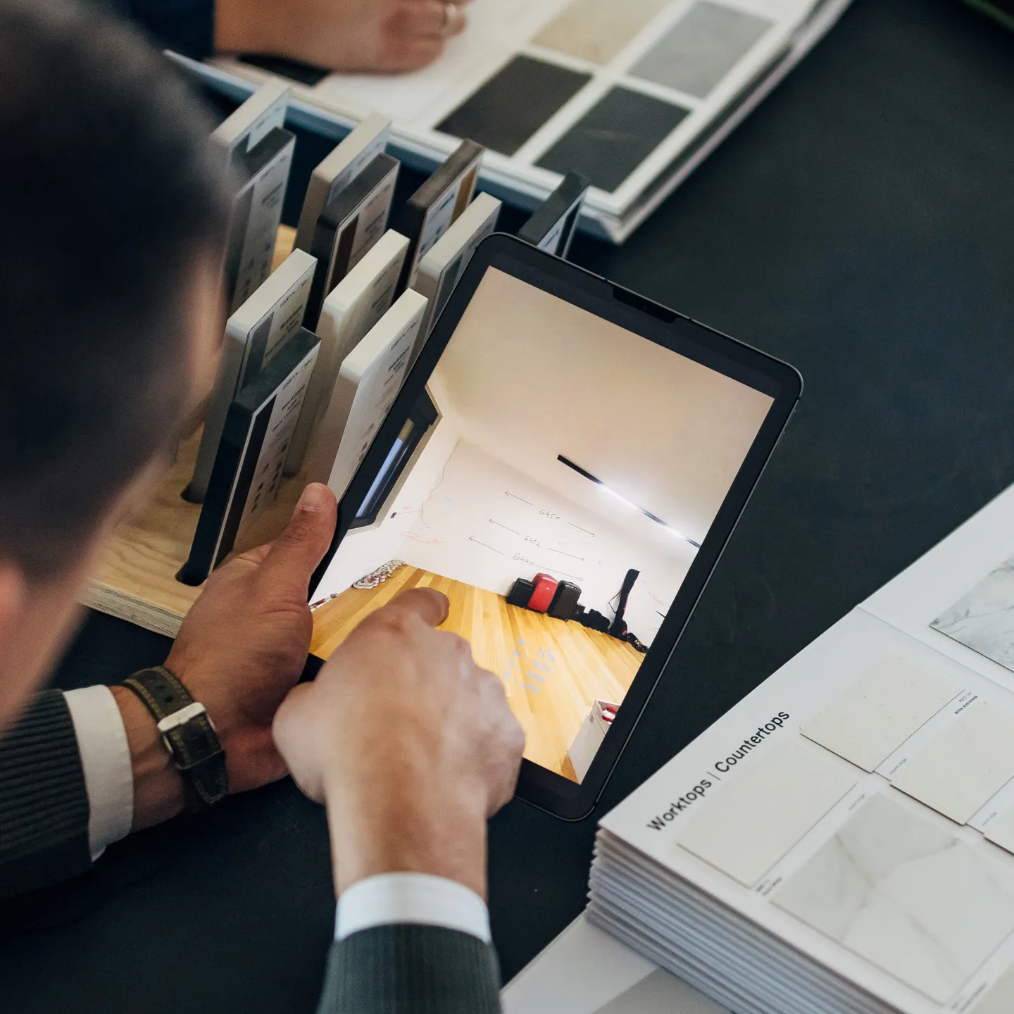 Osoba koristi tablet za pregled prostorije sa drvenim podom dok gleda uzorke radnih ploča na stolu.